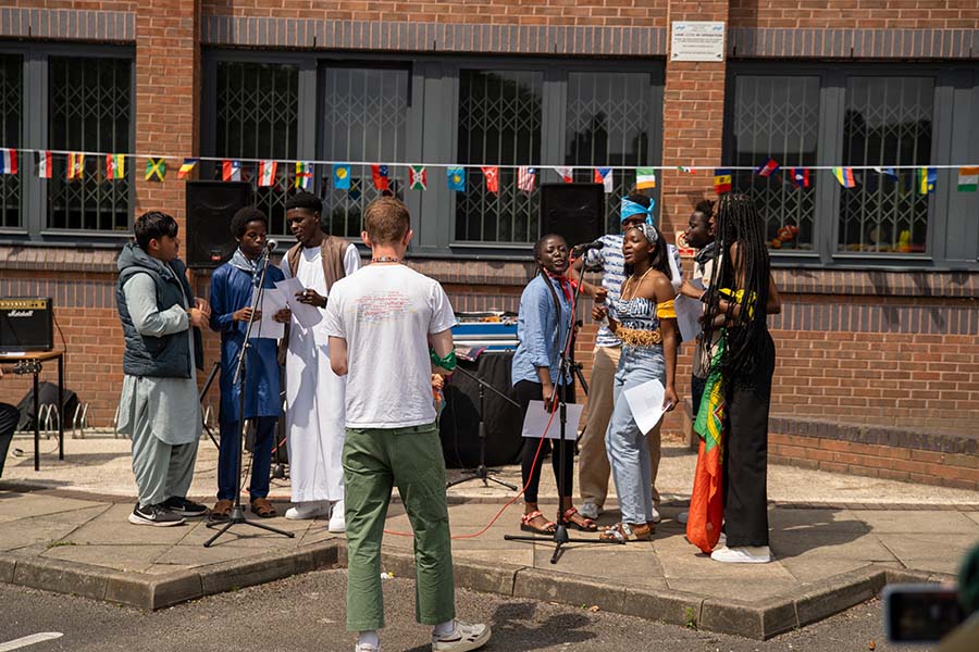 Two groups singing into microphones outside North Street building in the sun, next to colourful flag bunting for International Day 2024